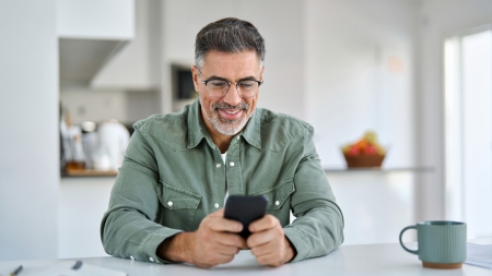 Ein Mann mit grauen Haaren und Brille lächelt, während er auf sein Smartphone schaut. Er sitzt an einem weißen Tisch in einem hell erleuchteten Raum mit einer modernen Küche im Hintergrund. Auf dem Tisch daneben steht eine blaugrüne Tasse.