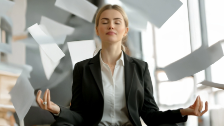 Eine Frau im Anzug sitzt im Schneidersitz in einer Meditationshaltung mit geschlossenen Augen und wirkt ruhig, während in einem Büro Papiere um sie herumfliegen.