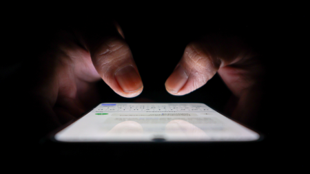 Nahaufnahme von Händen, die im Dunkeln auf einem Smartphone tippen, wobei der Bildschirm leuchtet und die Finger darüber beleuchtet werden. Das Telefon zeigt eine Messaging- oder Texting-App an.