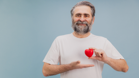 Ein älterer Mann mit grauem Haar und Bart lächelt, während er ein kleines rotes Herz in der einen Hand hält und mit der anderen darauf zustrebt.